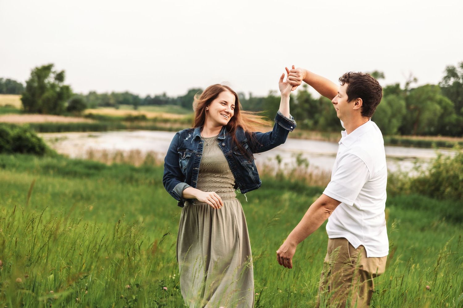 A couple dances together in a sunlit meadow near a pond while wearing casual summer attire.