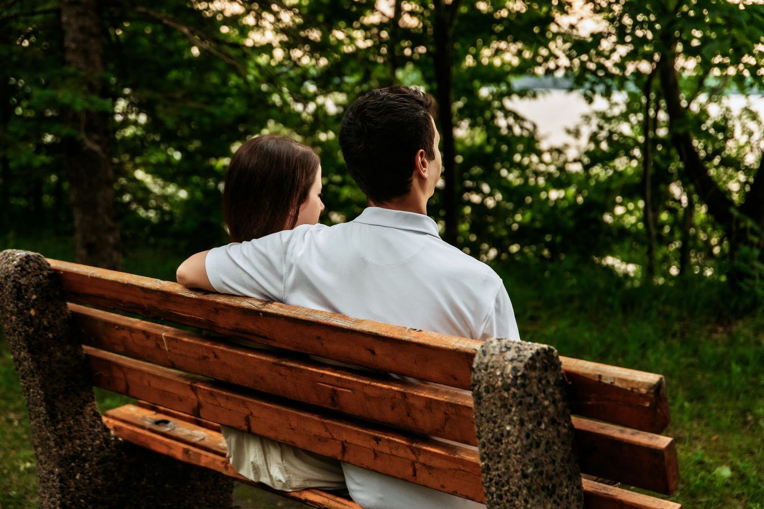 Two people sitting together on a wooden park bench surrounded by green trees at sunset.