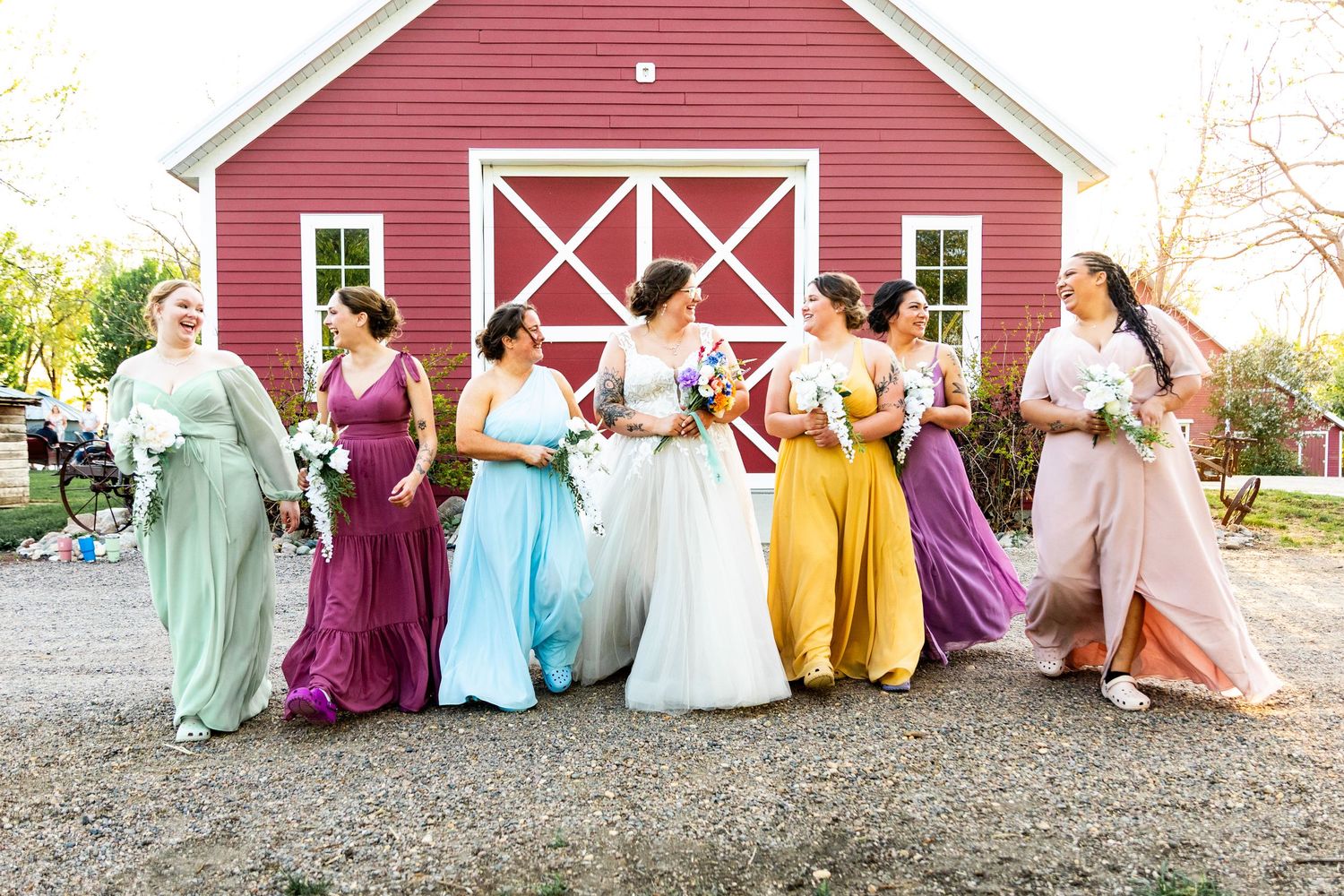 Bridal party in rainbow-colored dresses poses in front of red barn.