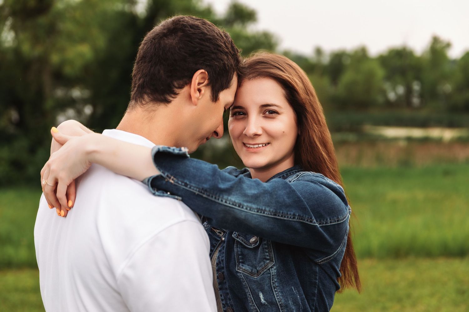 Young couple embracing outdoors with woman in denim jacket smiling at partner against green field background.