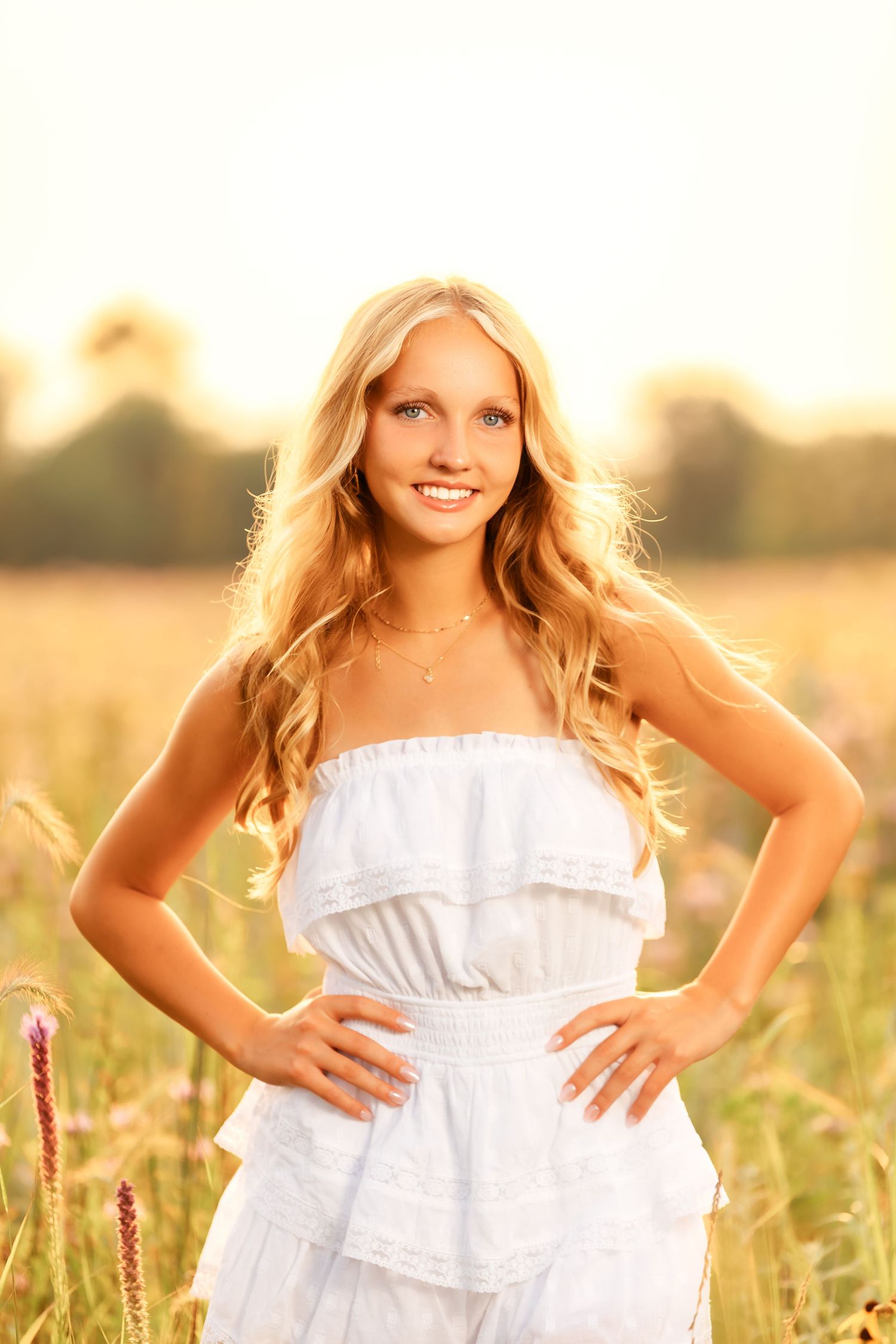 A person in a white summer dress poses in a sunlit field during golden hour with tall grass in the background.