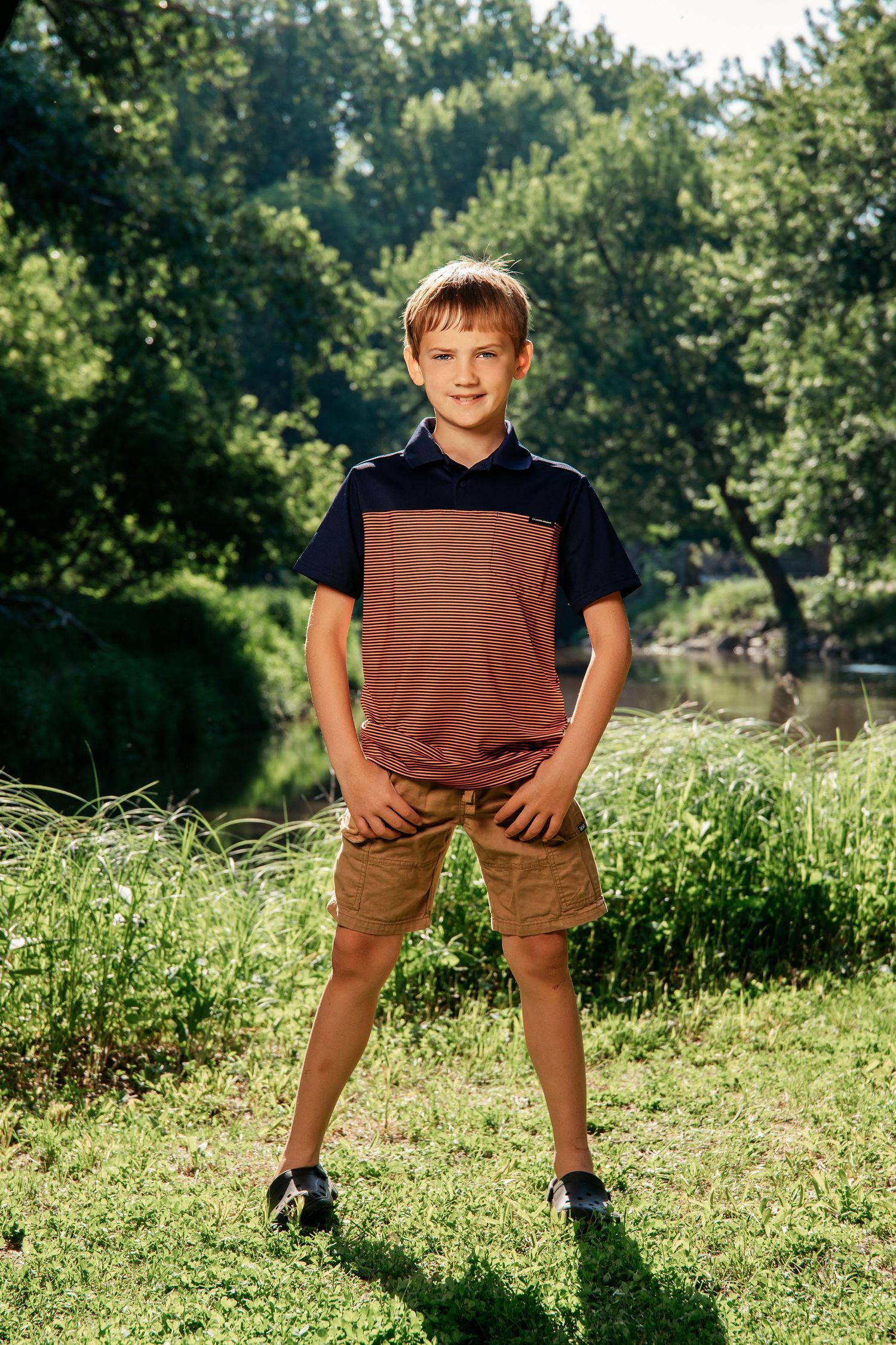 A youth in brown shorts and dark shirt stands casually in a grassy field during a bright summer day.