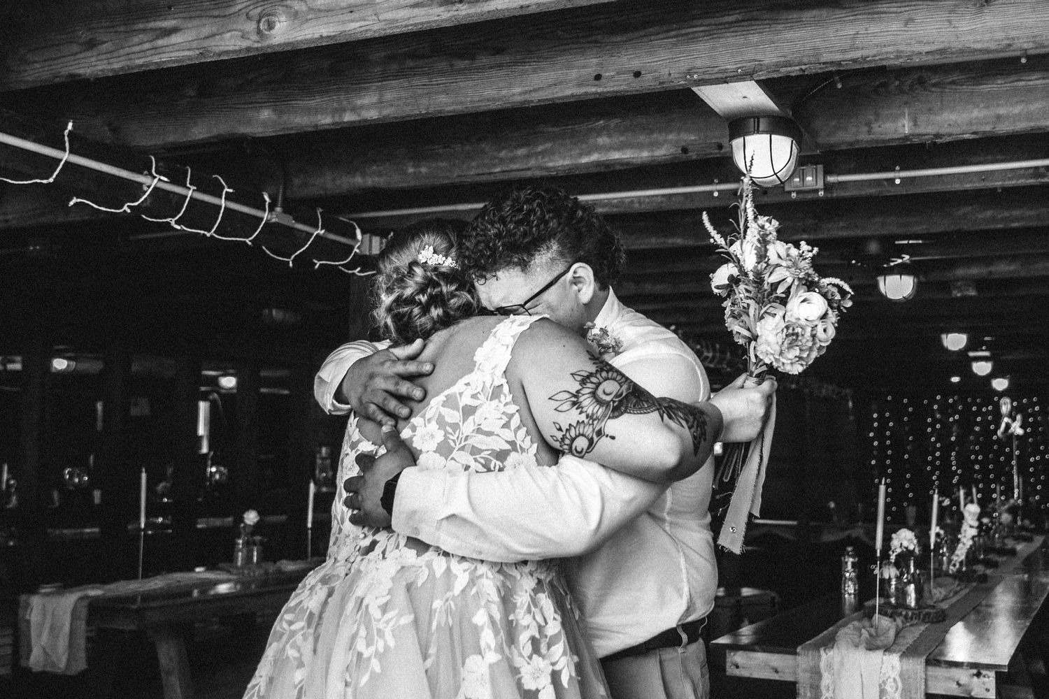 Black and white photo of wedding guests dancing in a rustic barn venue with overhead lighting.
