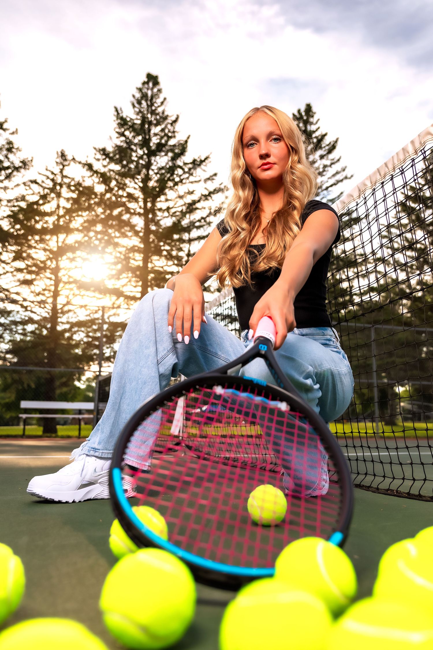 Athletic woman in casual attire posing with tennis equipment and neon yellow balls on an outdoor court at sunset.
