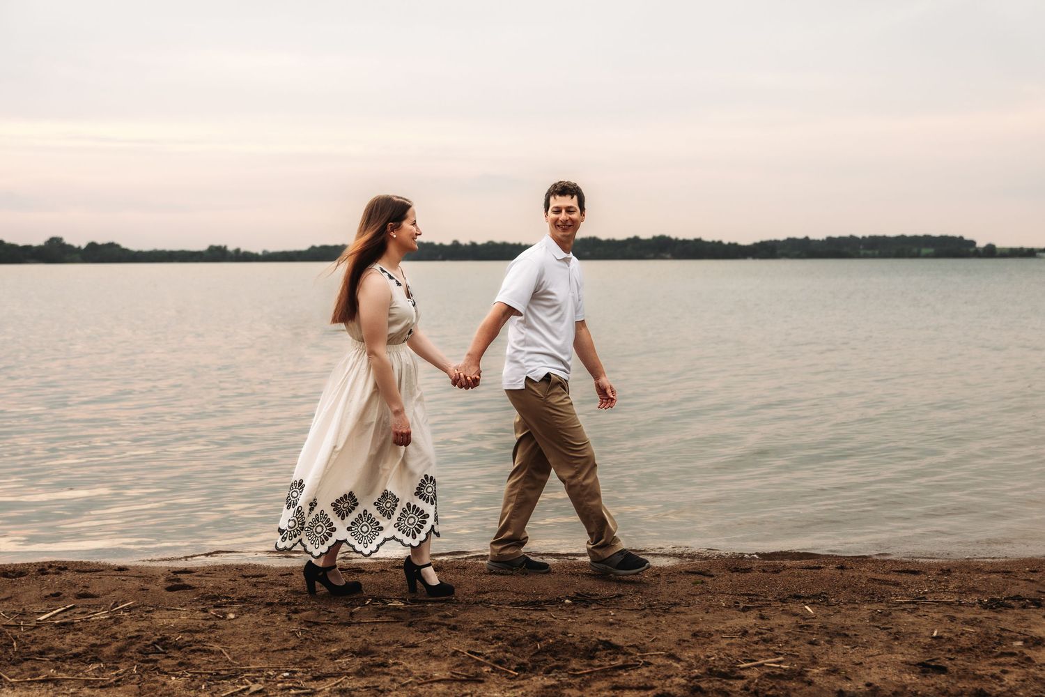 Couple walking hand in hand along shoreline at sunset wearing casual summer attire.