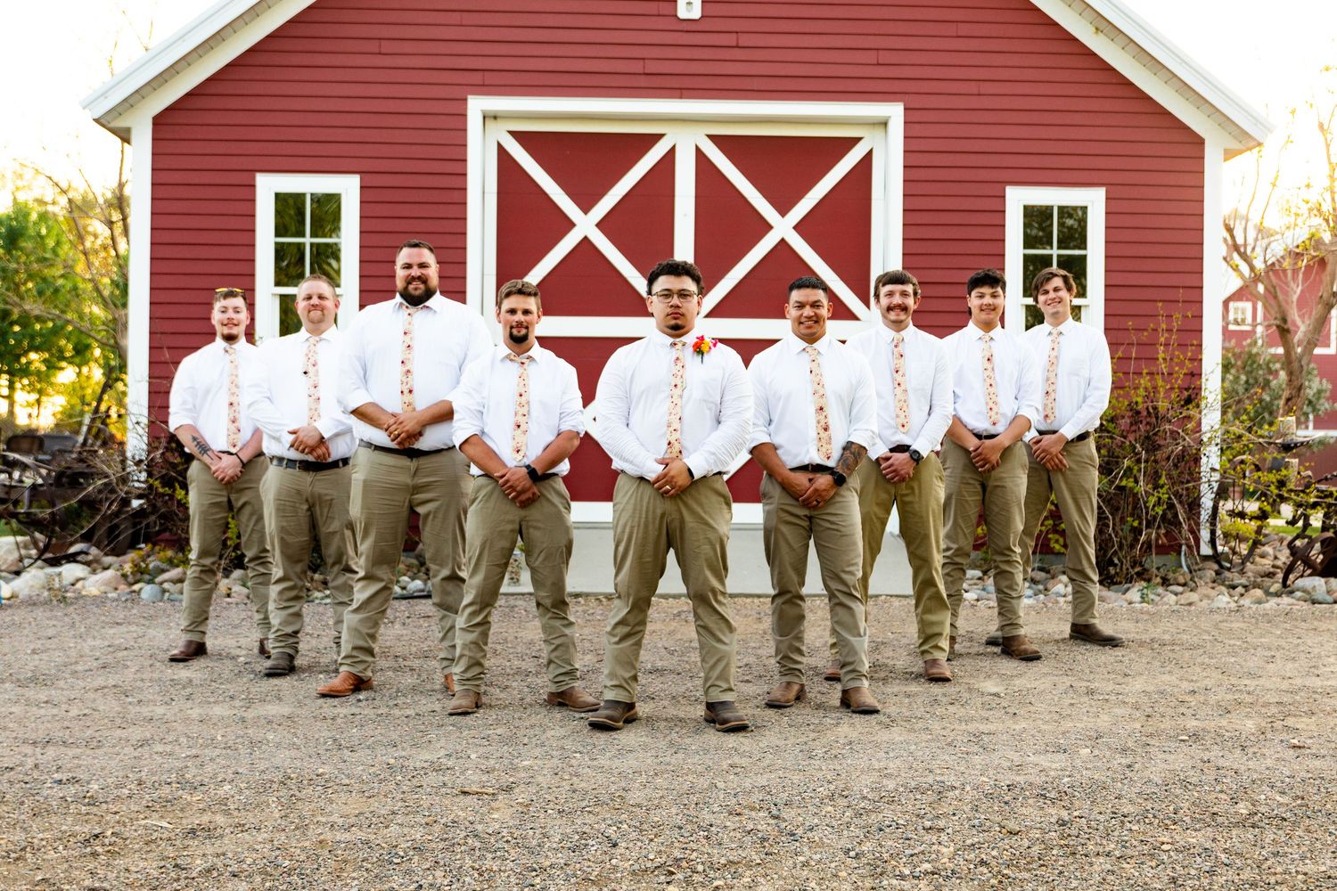 Groomsmen in khaki pants and white shirts pose in a line in front of a red barn.