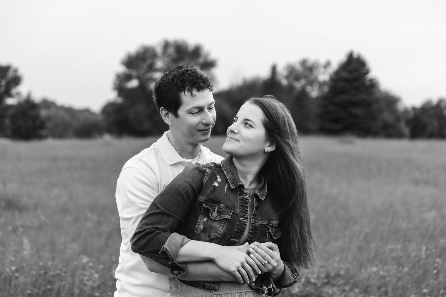 A couple shares a tender moment together with their black dog in a black and white photograph taken in a meadow.