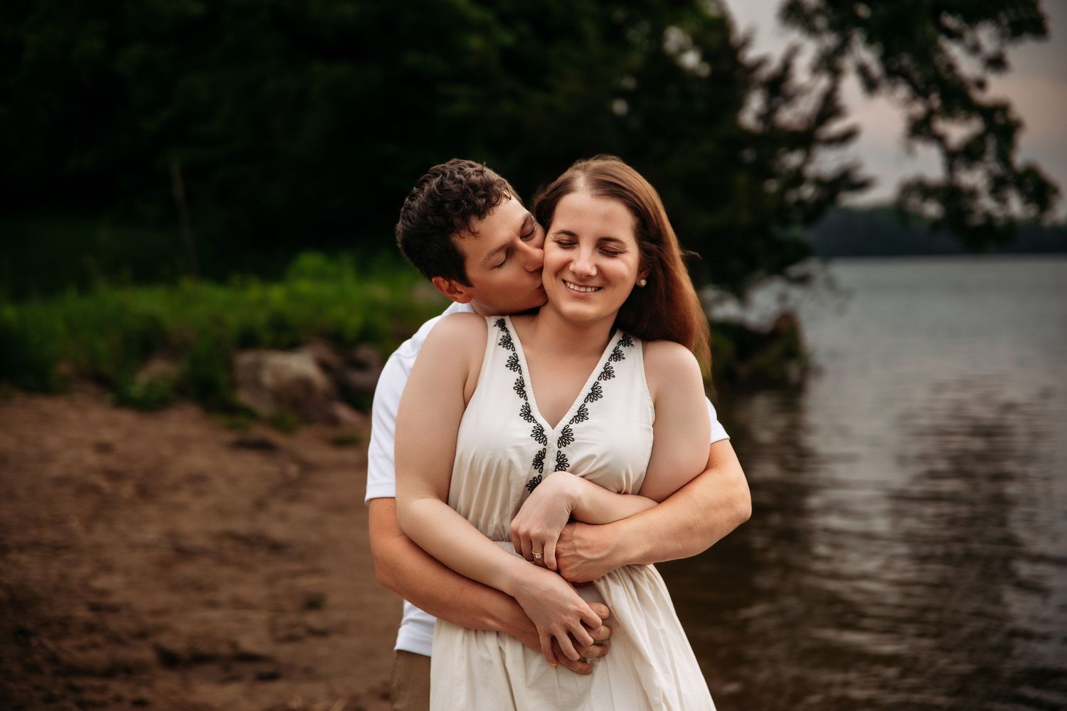 A romantic couple embraces while wearing white dresses on a serene beach at sunset near calm waters.