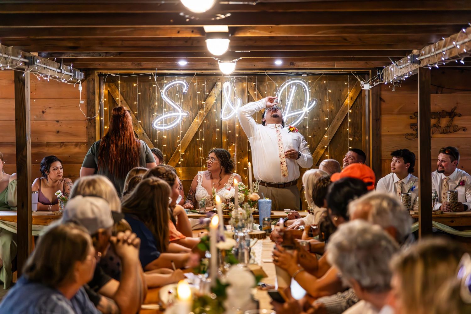 Wedding reception guests celebrate inside a rustic wooden venue with decorative lighting.