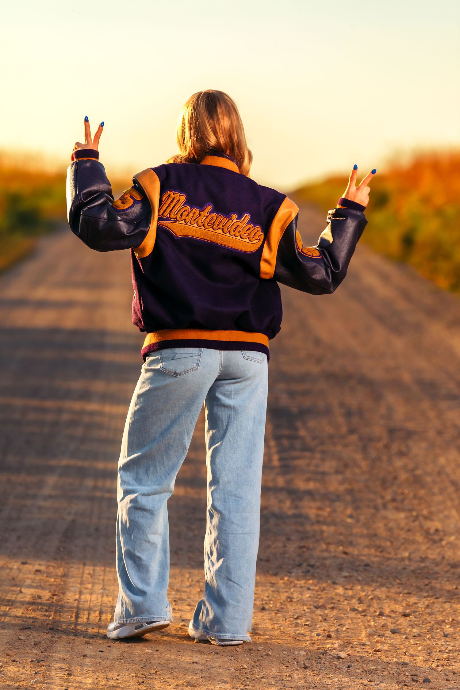 A figure wearing a varsity jacket and jeans gives peace signs on a dirt road at golden hour.