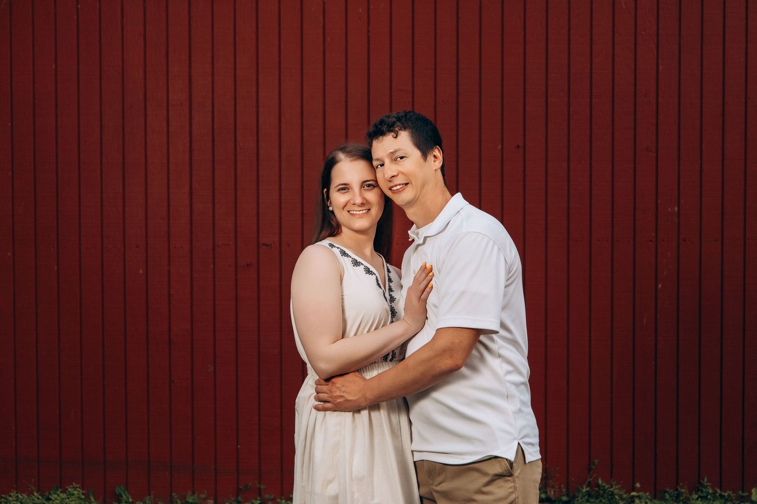 A couple in white outfits pose together against a red wooden wall background during a romantic photoshoot.