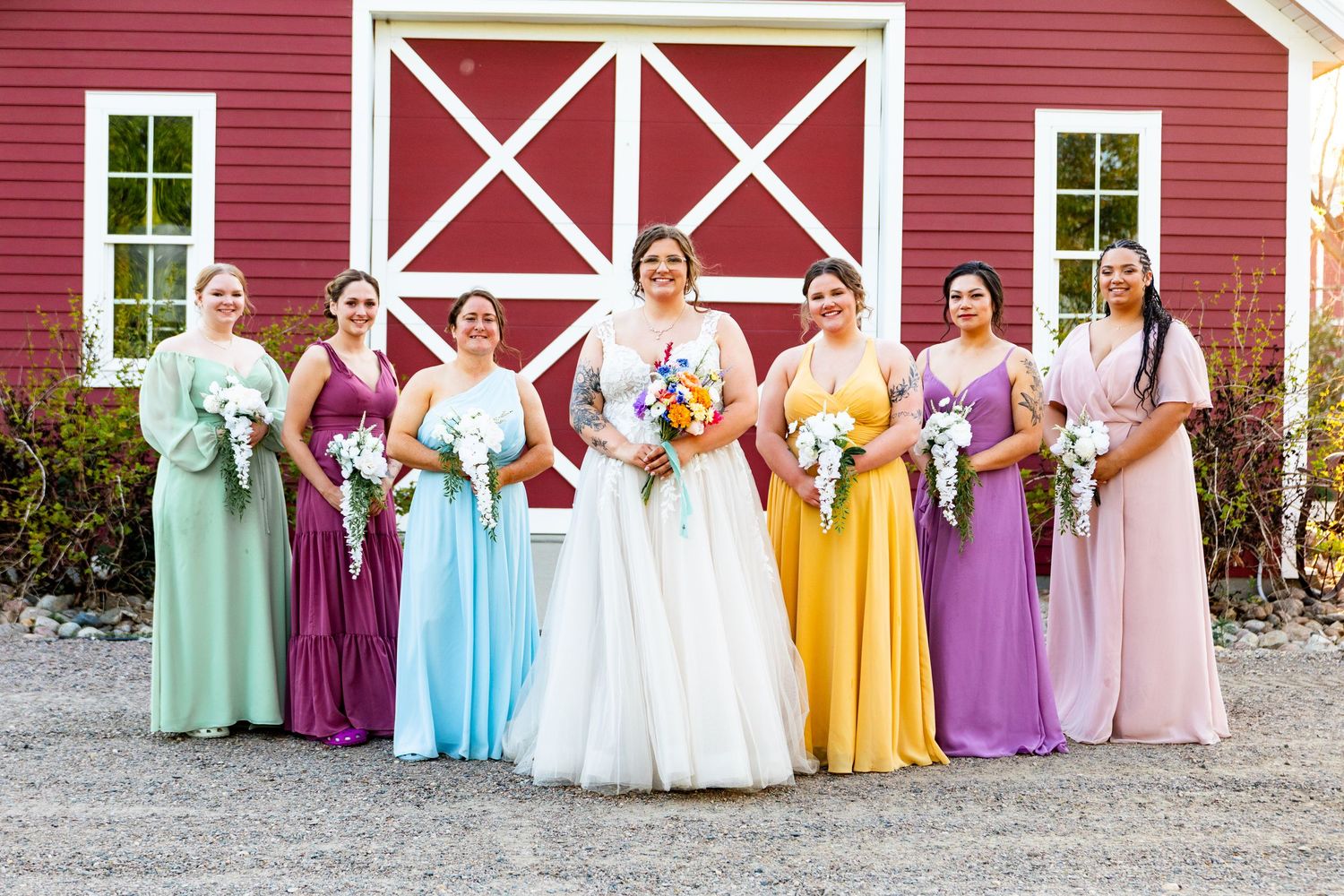 Wedding party poses in colorful pastel dresses against classic red barn backdrop.