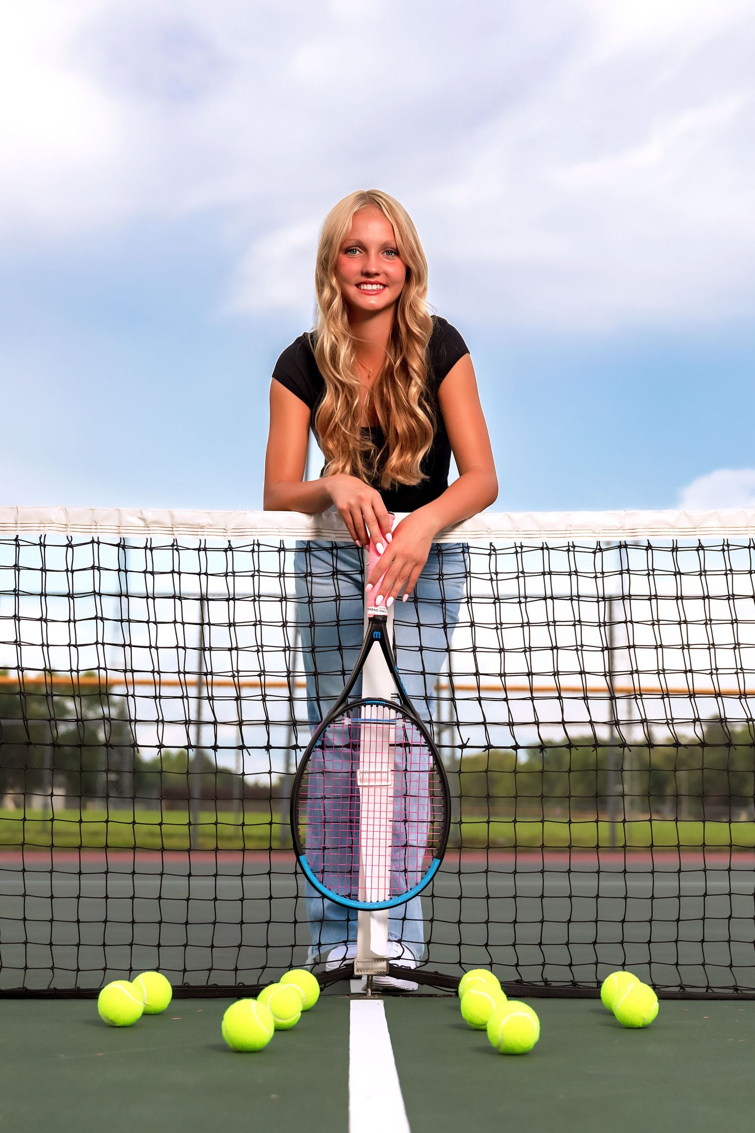 Tennis player poses with racket and tennis balls on court against blue sky and net backdrop.
