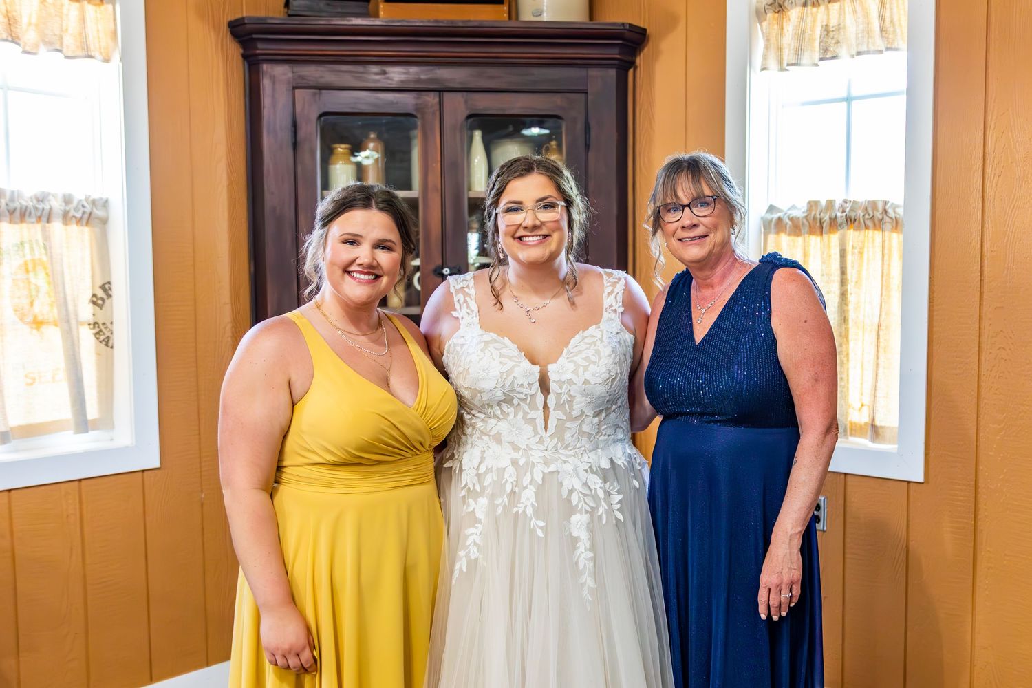 Three women in formal dresses of yellow, white, and navy blue pose together by a window in an interior room.