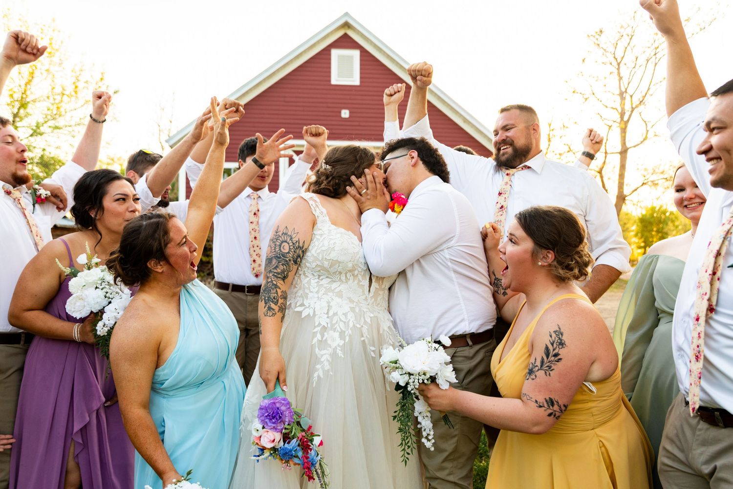 Wedding party celebrates with enthusiasm outside red barn during golden hour.