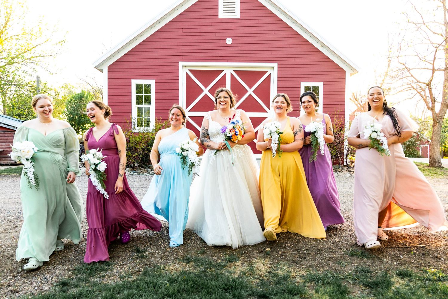 Wedding party wearing rainbow colored dresses line up against a red barn backdrop.