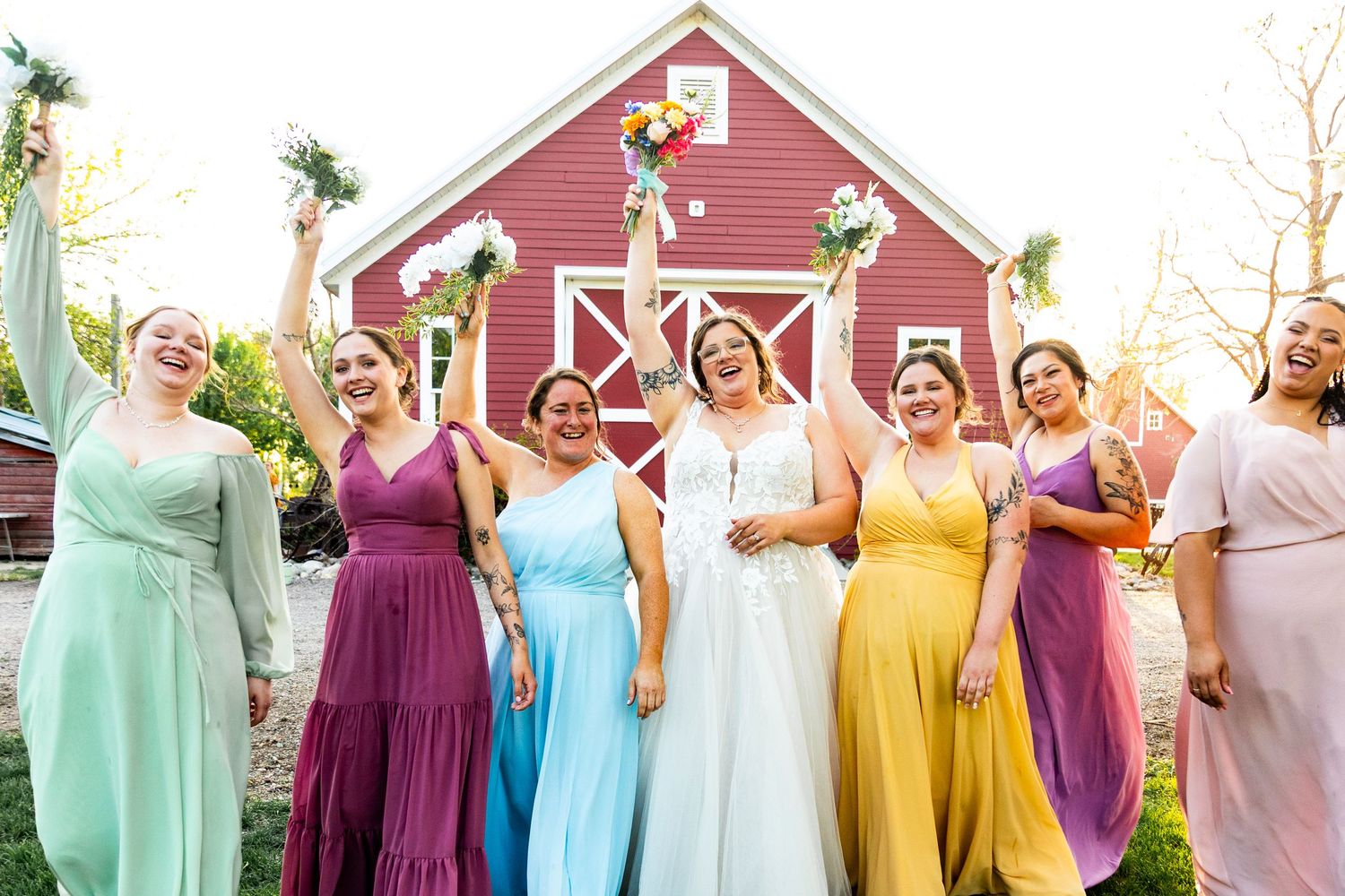 Wedding party members in pastel rainbow dresses lined up against a rustic red barn.