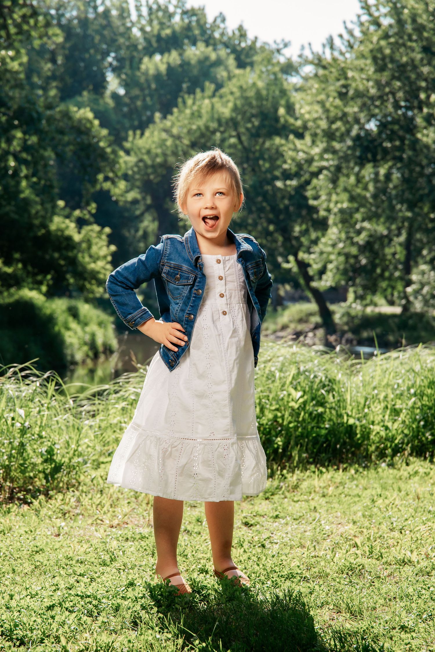 A young person in a white dress and denim jacket plays joyfully in a sunny grassy field on a summer day.