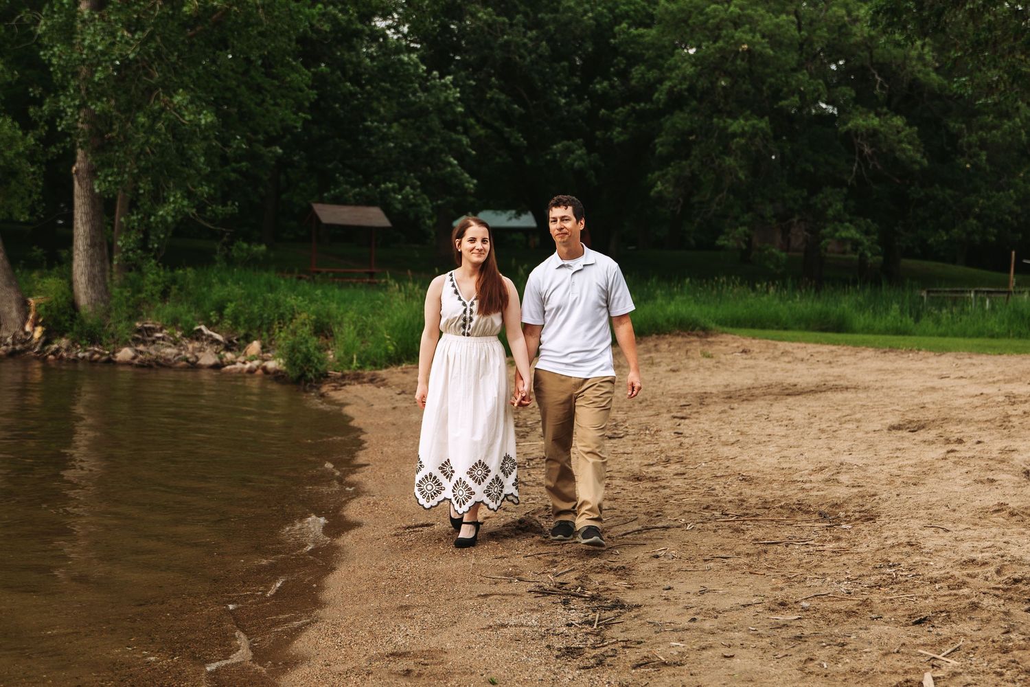 A couple in summer attire walks hand in hand along a sandy beach by a wooded shoreline.