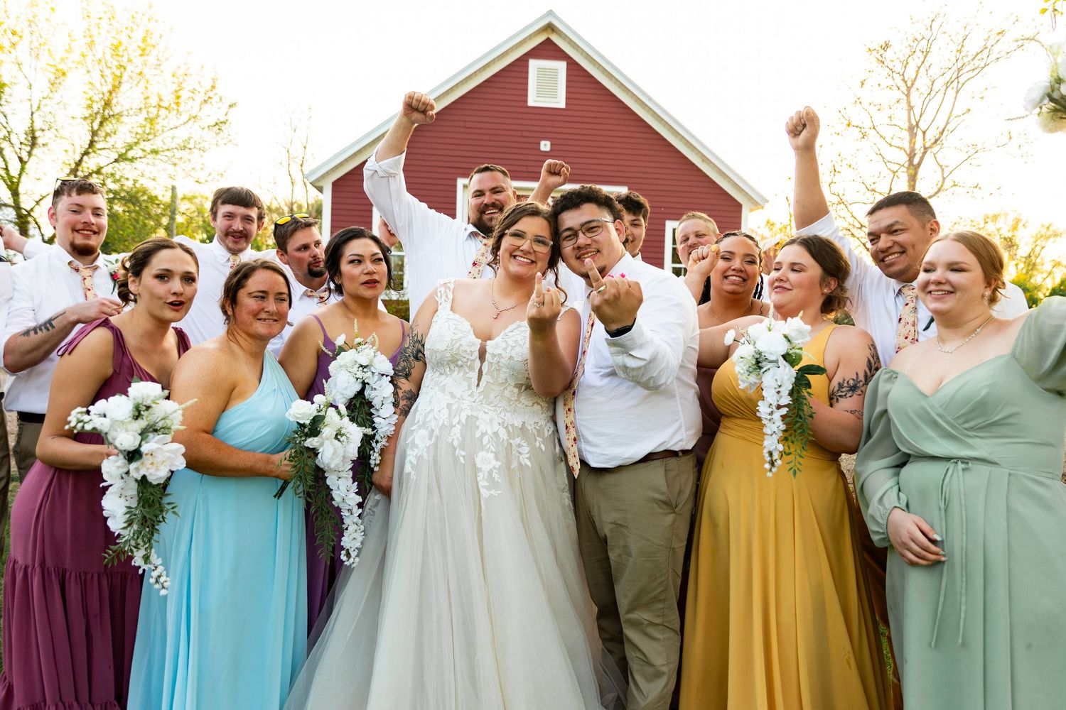 Wedding party in colorful pastel dresses poses joyfully in front of red barn.