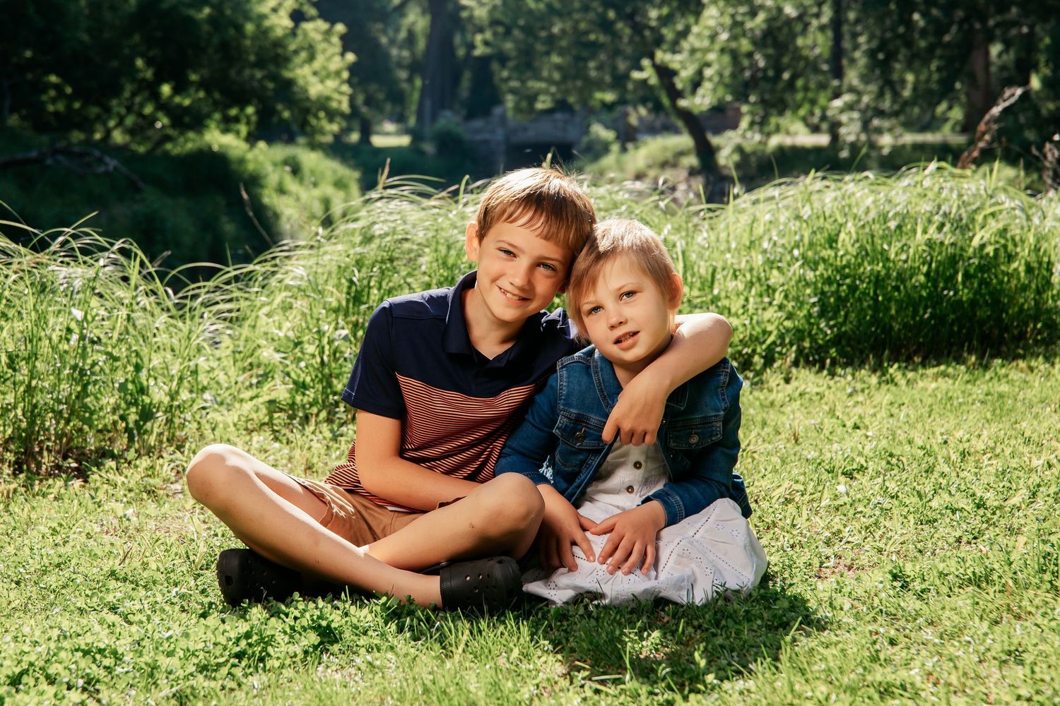 Two children sit close together on grass in a sunny park, sharing a warm moment in the natural setting.