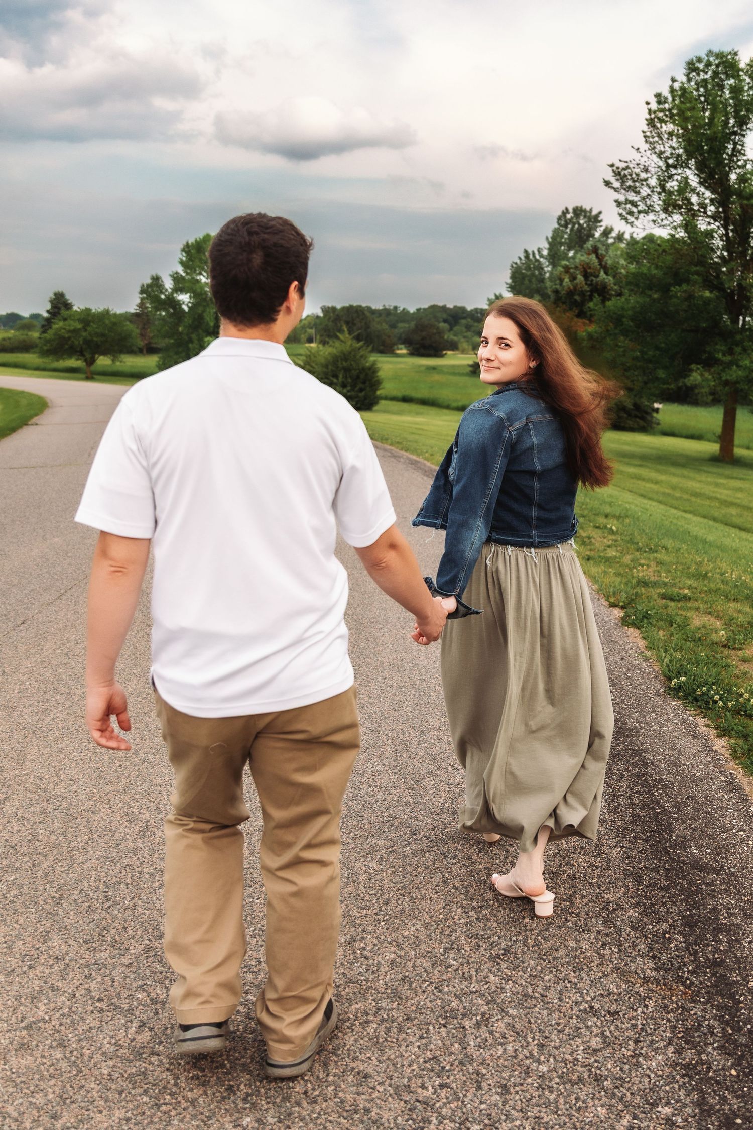 A couple walks hand in hand along a winding path through a green park on a cloudy summer day.