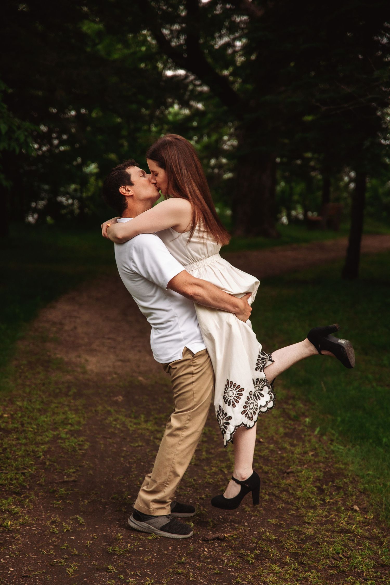 A couple embraces playfully on a forest path while the woman lifts her foot.