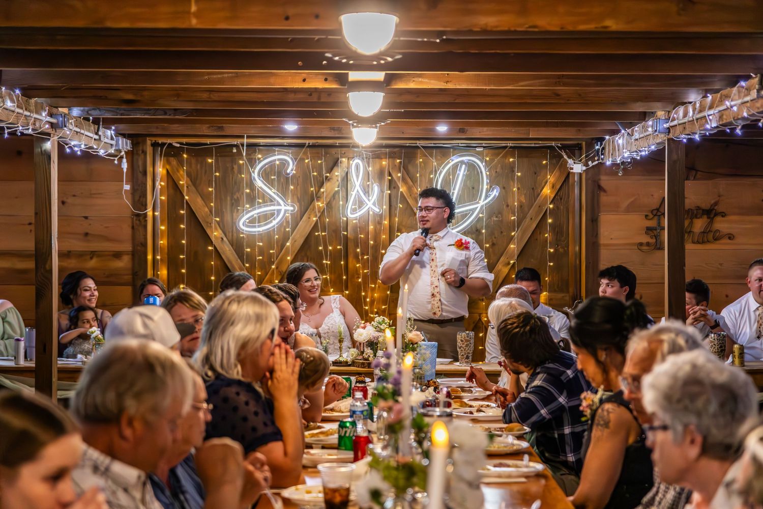 Guests enjoy dinner at a cozy indoor wedding reception with exposed wooden beams and string lights.