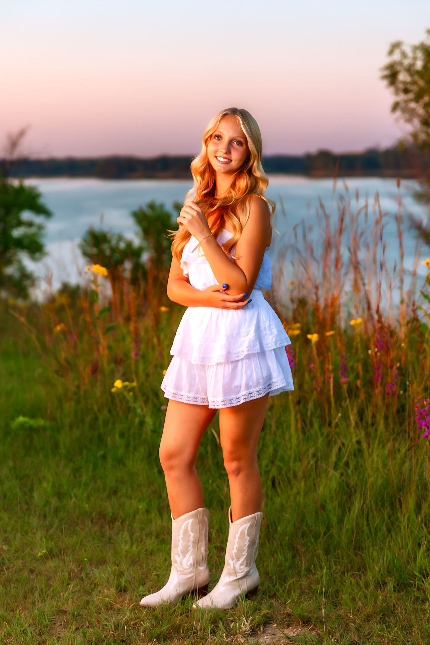 Woman in white dress and cowboy boots stands in field by lake during sunset.