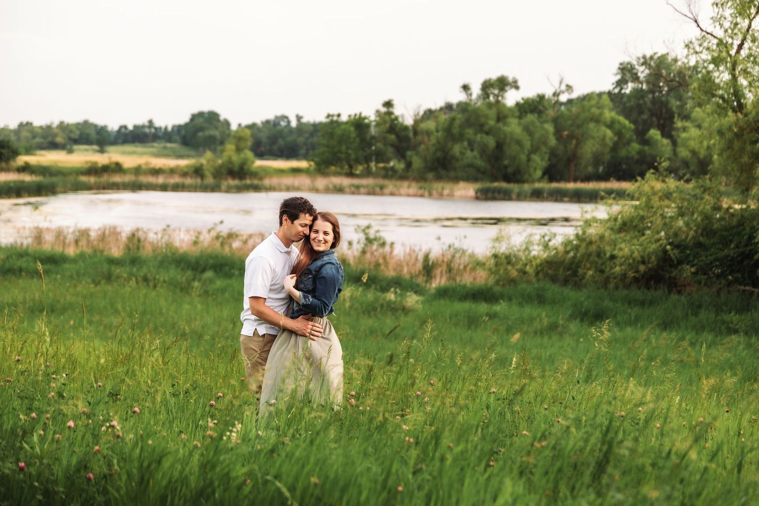 Couple romantically embracing in a sunny green meadow with golden light filtering through trees.