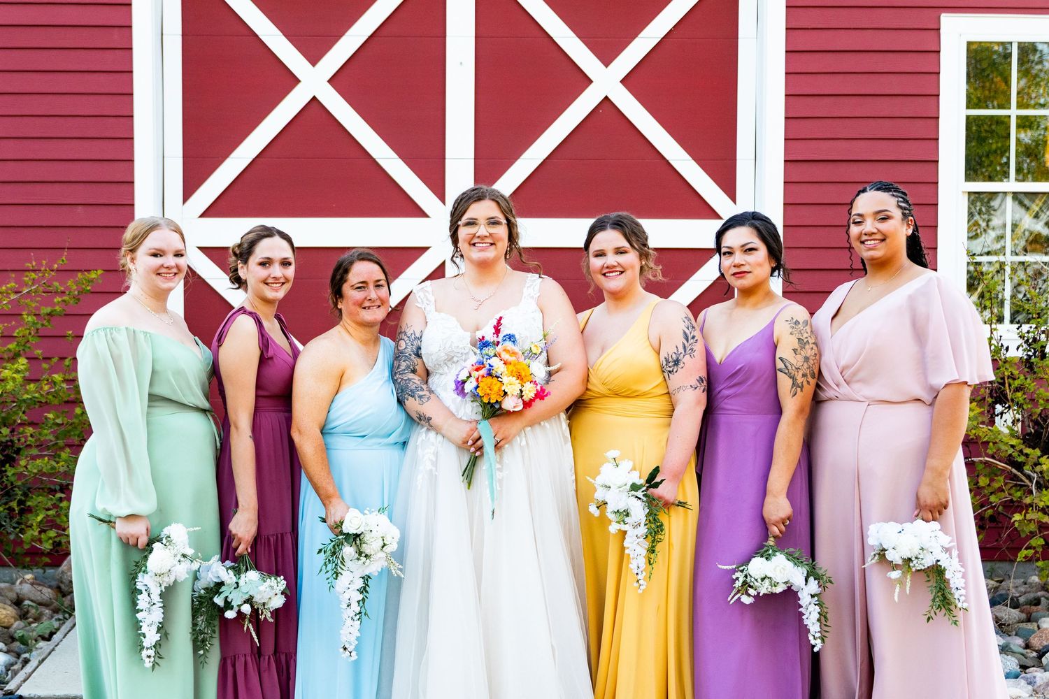 Bridal party in pastel rainbow dresses poses in front of red barn with white trim.