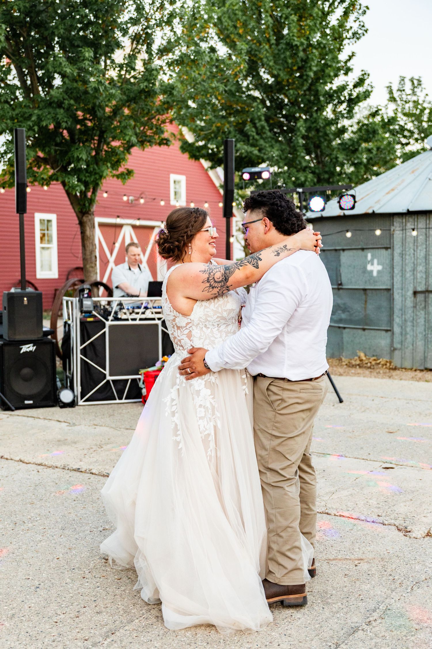 A newlywed couple shares their first dance outdoors near a red barn and DJ setup.