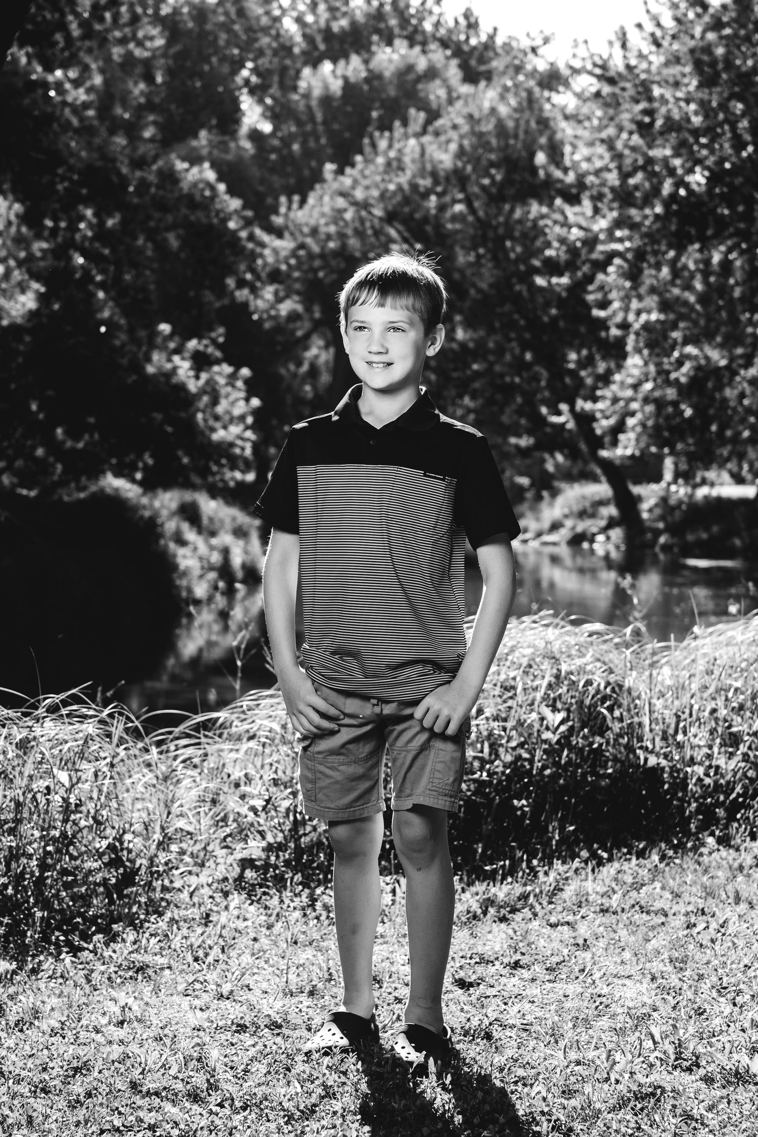 Black and white portrait of a young boy in casual attire standing outdoors on a sunny day.
