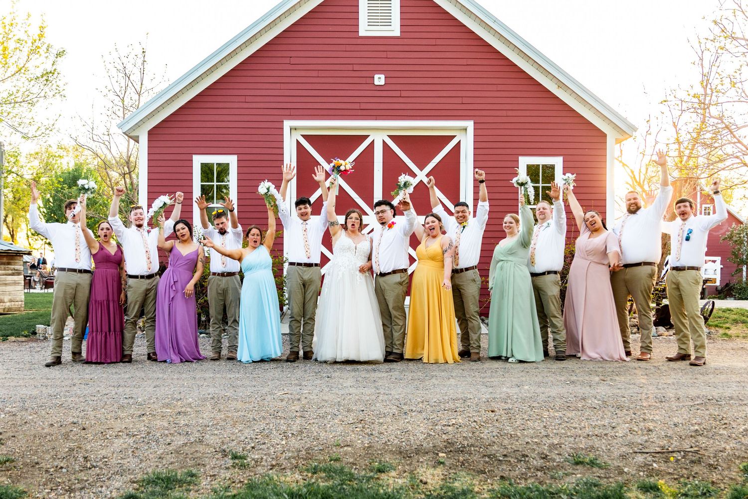 Colorful wedding party wearing rainbow pastel dresses posed in front of a red barn.