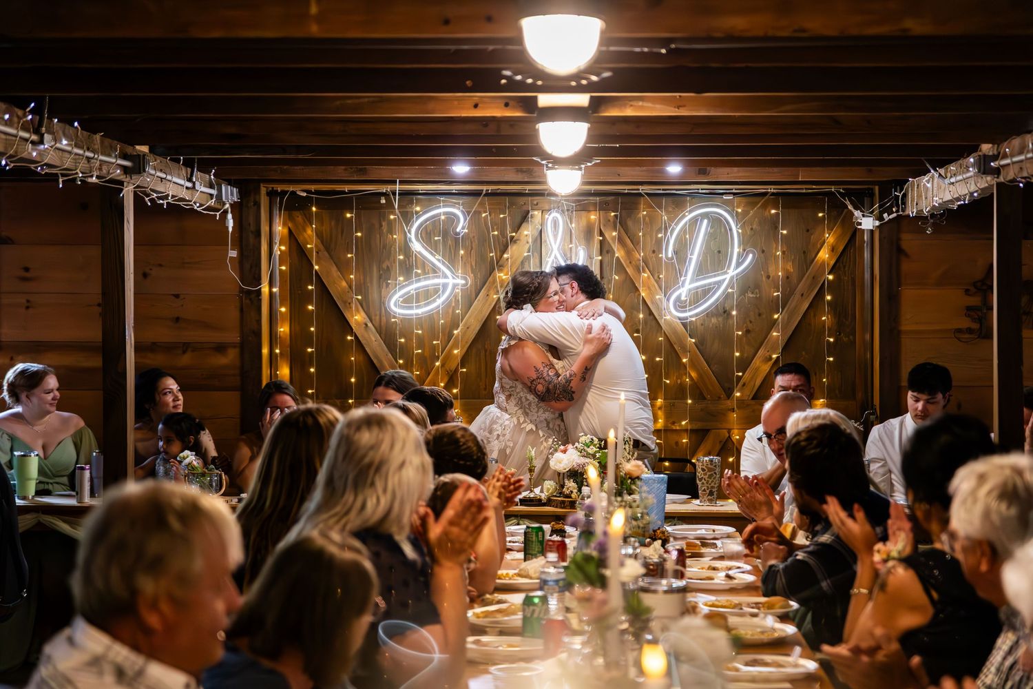 Indoor barn wedding reception with hanging lights and guests seated at long tables.