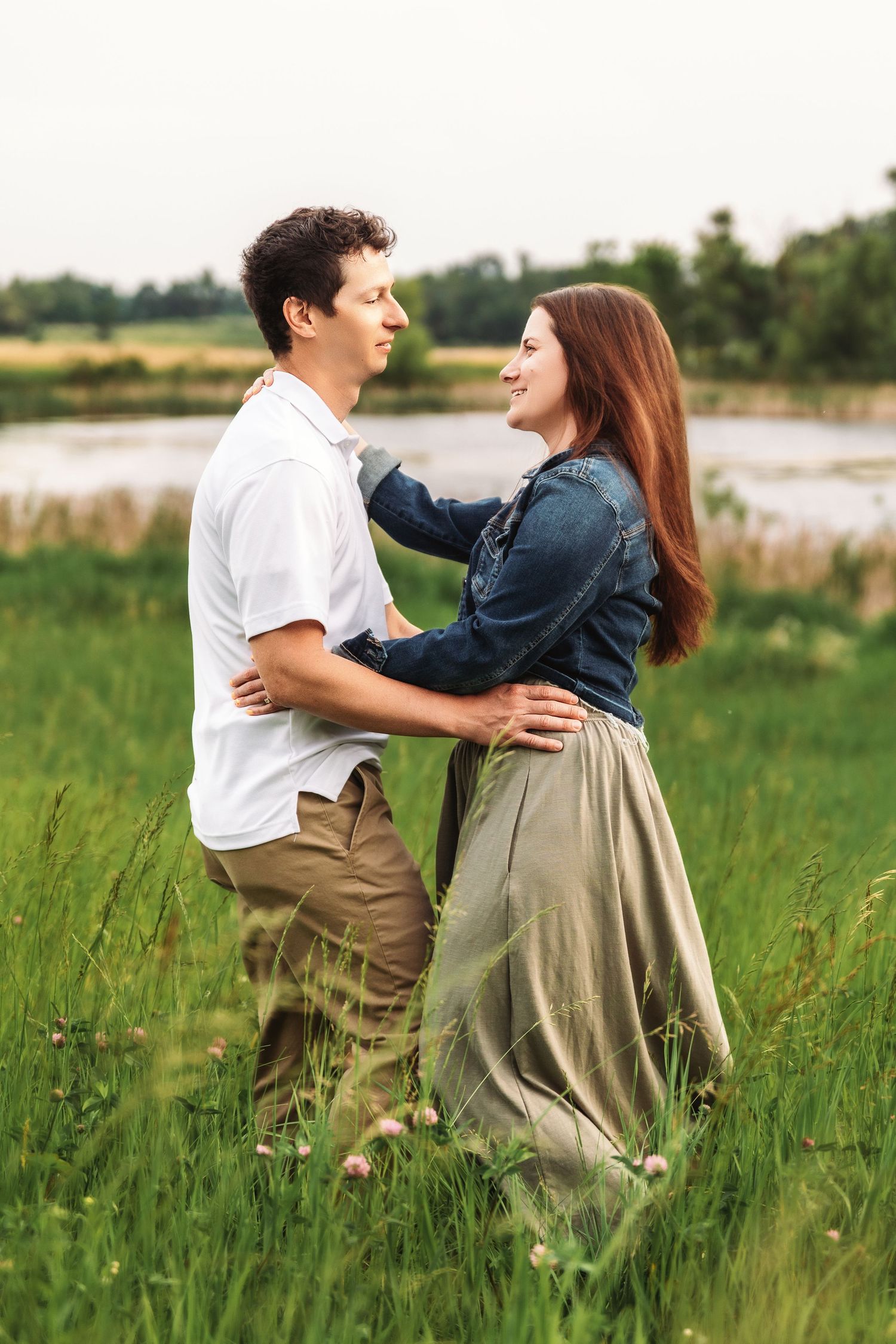 A romantic couple embraces in a field of tall grass with a lake in the background.