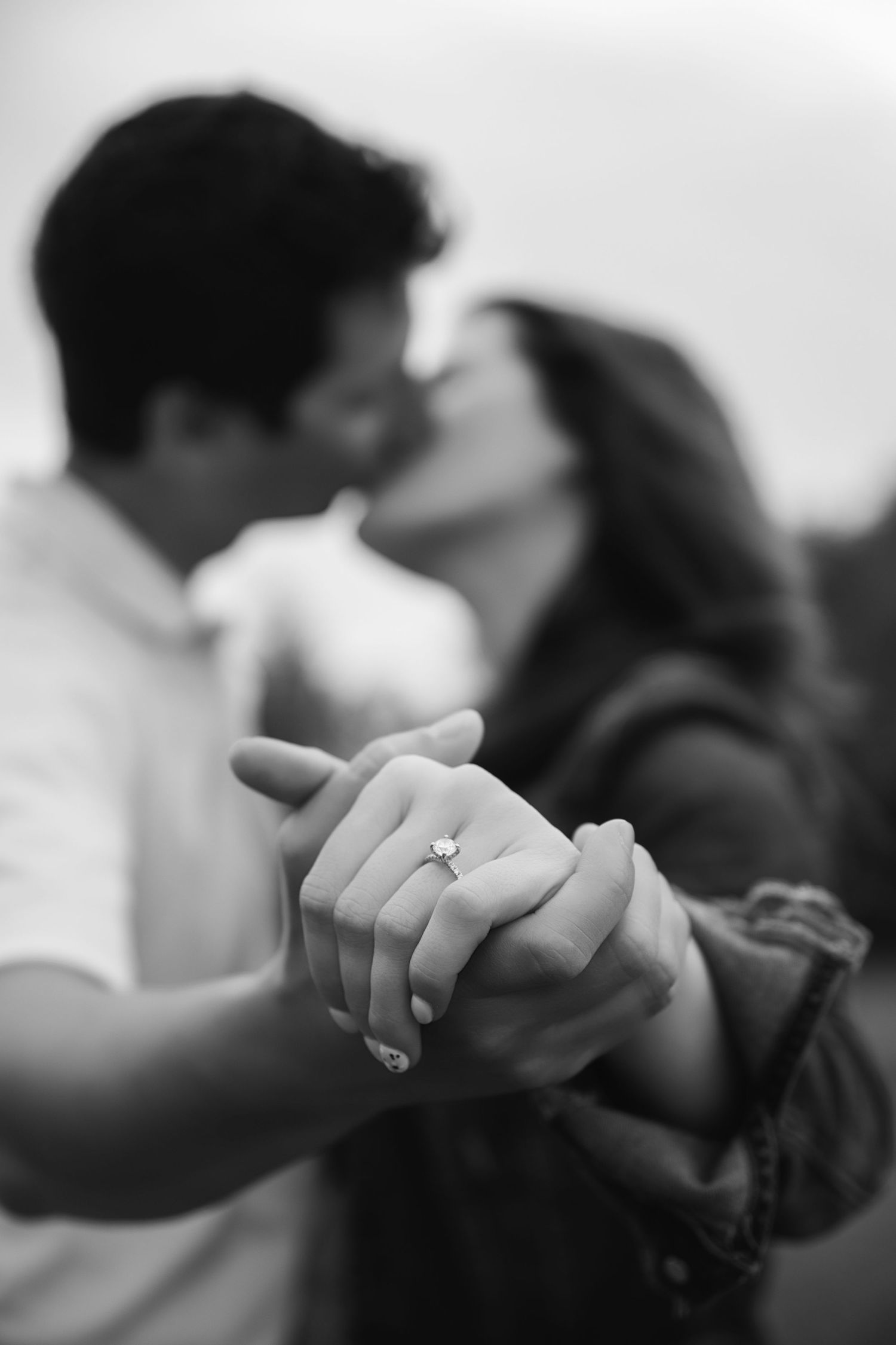 Close-up black and white shot of clasped hands with an engagement ring, with a romantic couple kissing in the background.