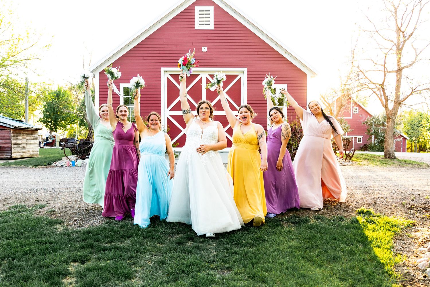 Wedding party in colorful dresses poses in front of a classic red barn.