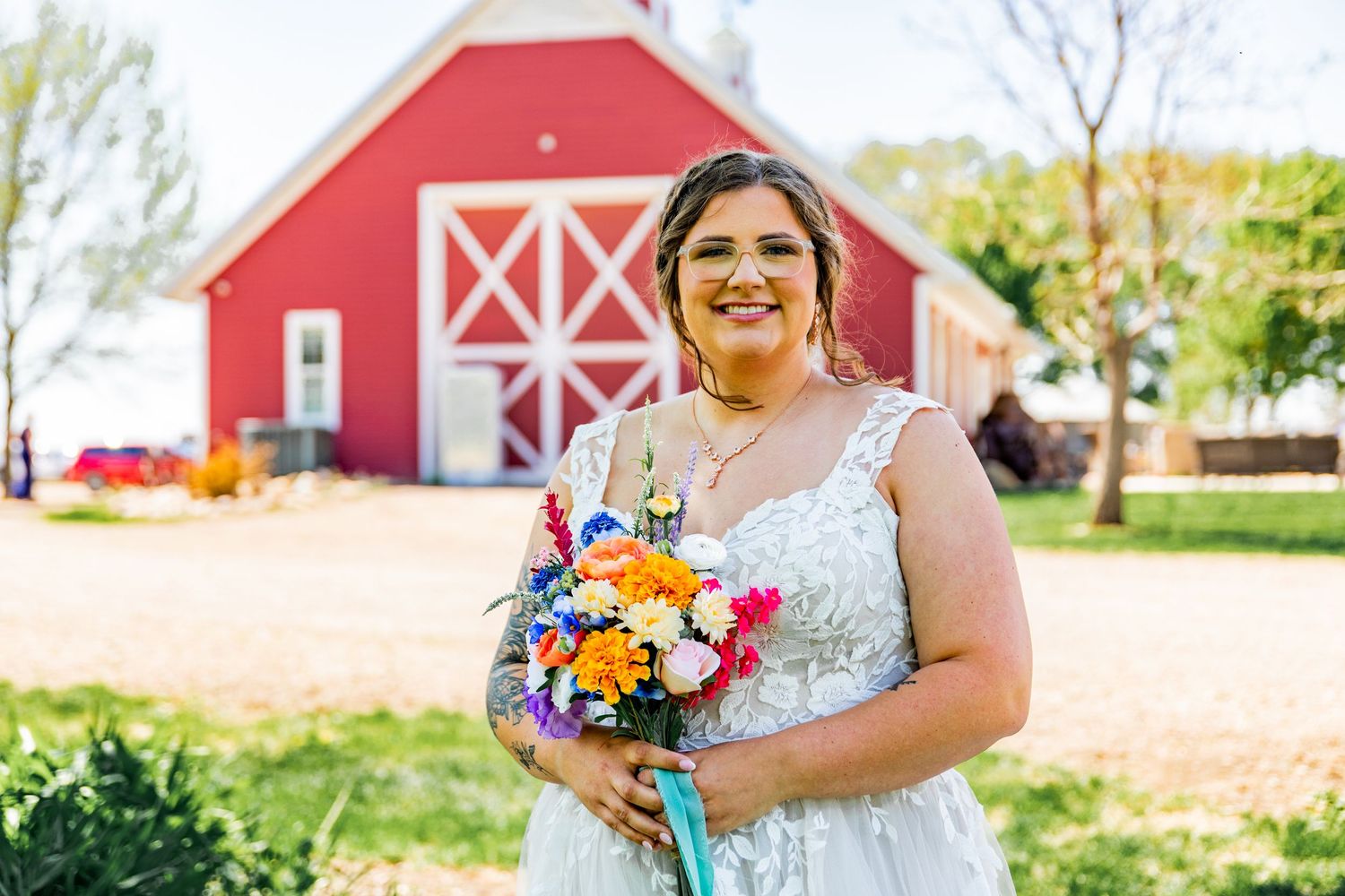 Bride holding vibrant bouquet poses in front of red barn on sunny wedding day.