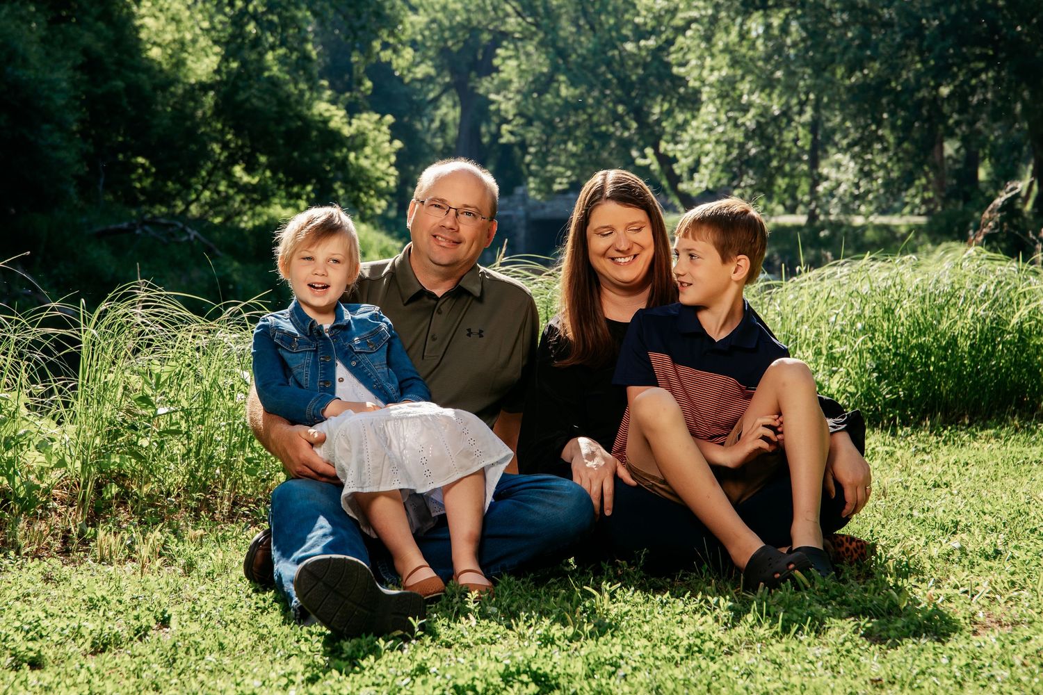 Family of four sitting together outdoors on green grass with sunlit trees in background during summer day.
