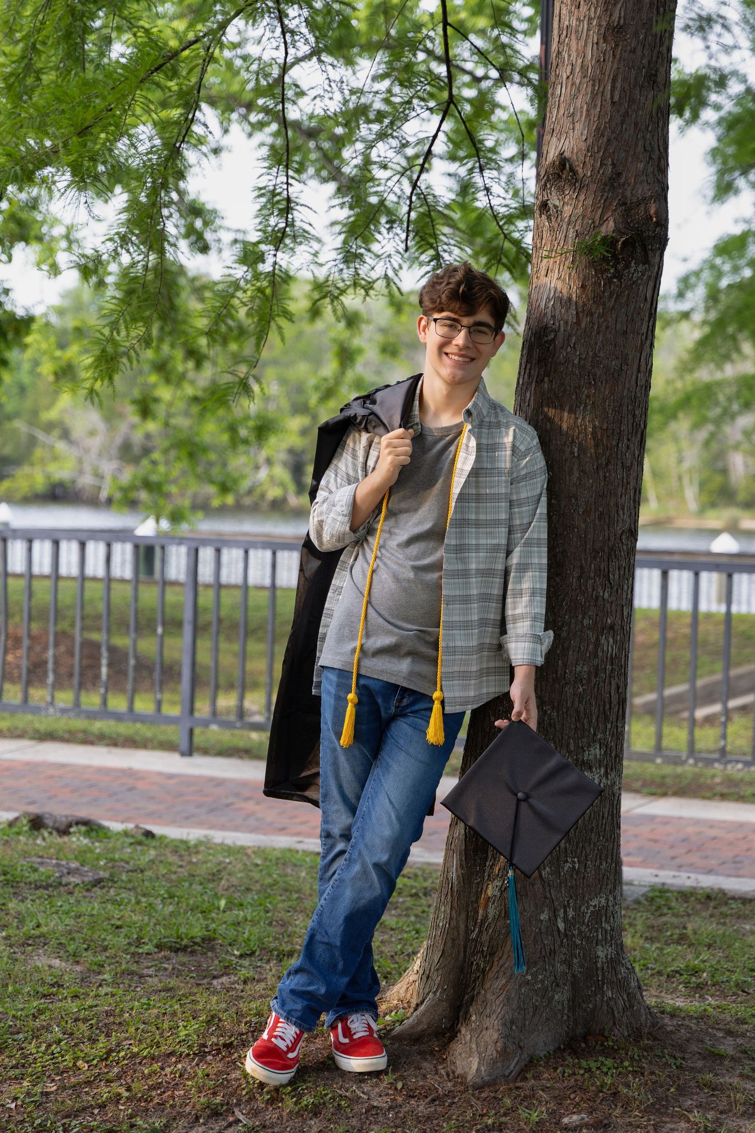 Senior portrait Class of 2025—handsome young man casually leaning on a tree with cap & gown at Milton, FL, Photography by Rachel.