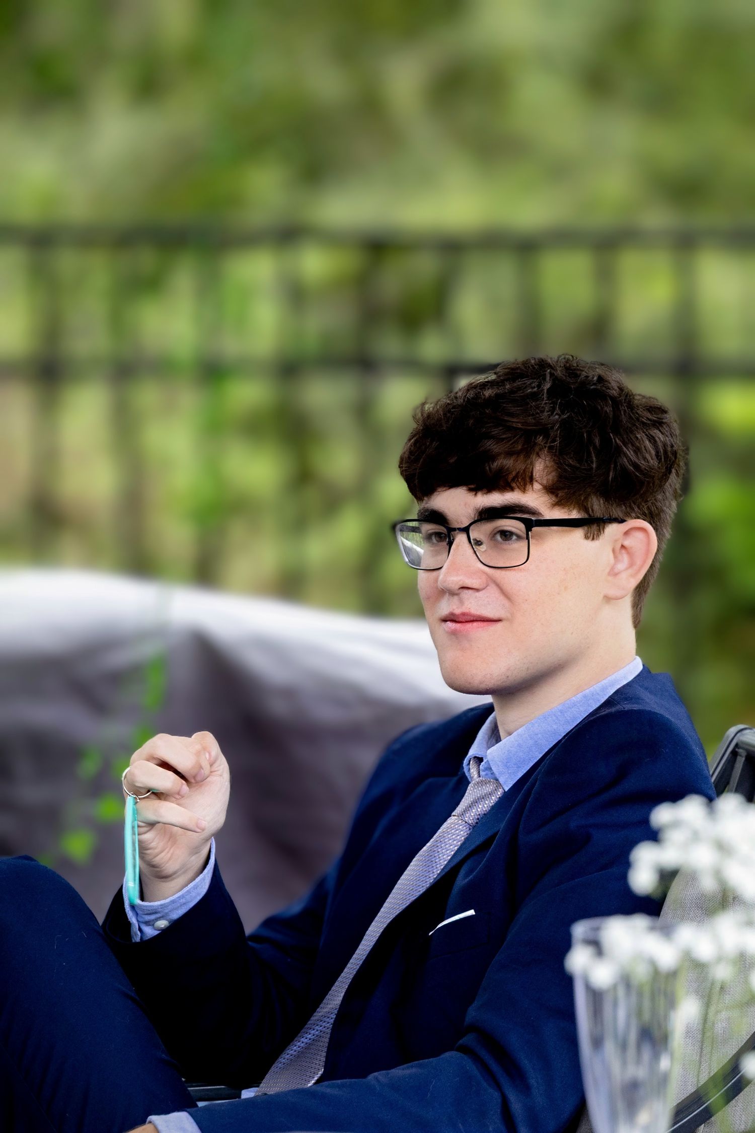 Candid natural light senior portrait in Pace, FL—handsome young man in a blue suit, seated at outdoor event table. Photography by Rachel.