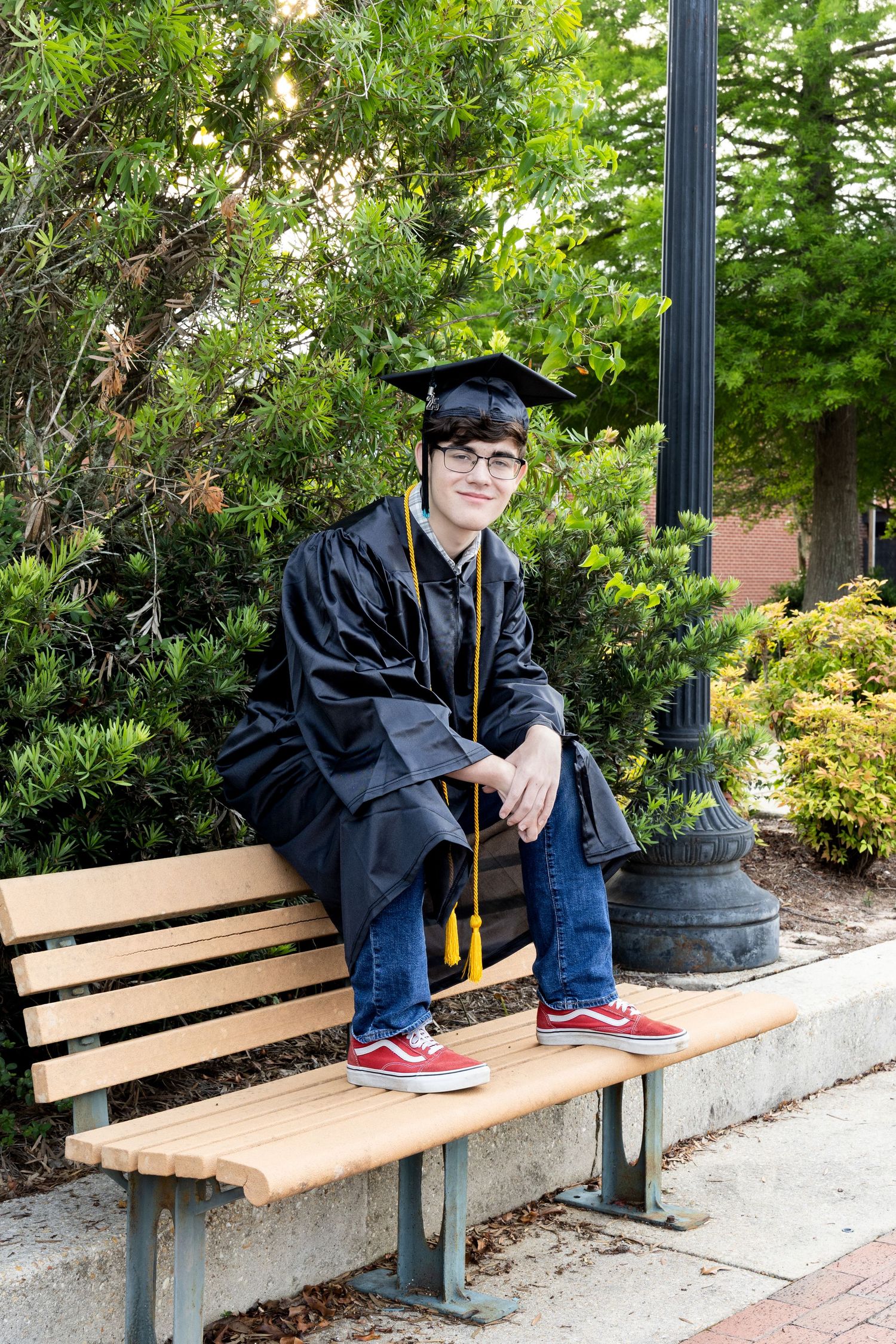 Class of 2025 senior in cap & gown, relaxed pose on back of park bench at Milton Riverwalk—friendly, handsome, full of personality.