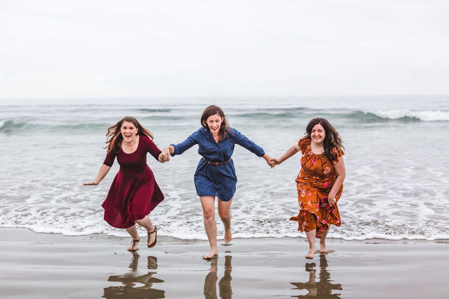 Three friends in colorful dresses jumping together on a misty beach.