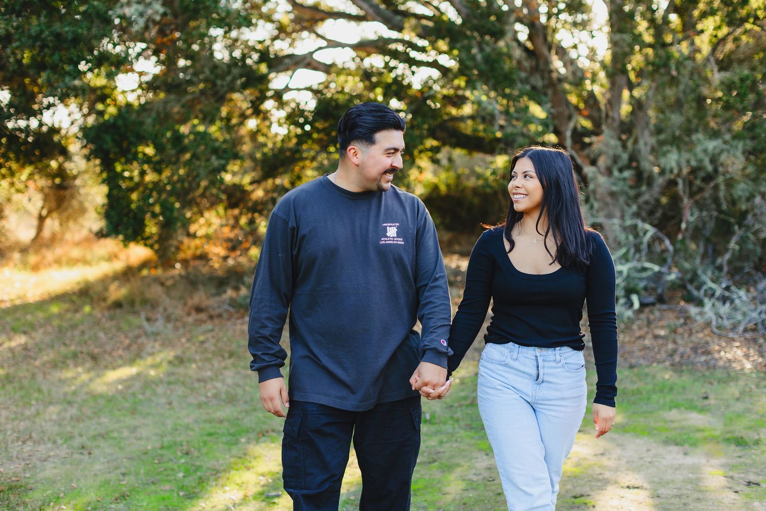 A couple wearing casual dark jackets walks together in an outdoor setting with trees and foliage in the background.