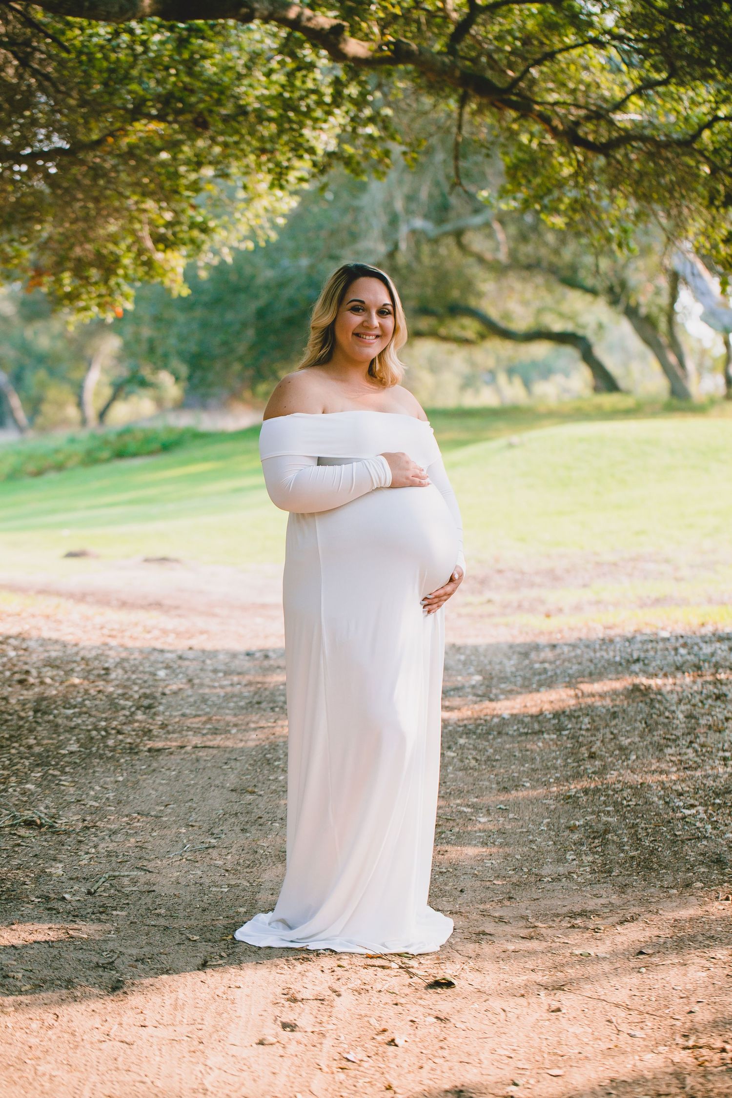 Expectant mother in white off-shoulder dress poses gracefully under tree canopy.