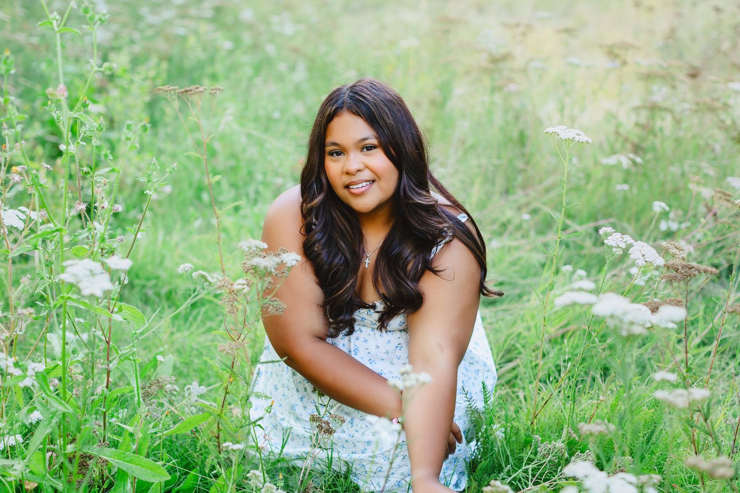 A person in a white floral dress sits smiling in a meadow of tall grass and wildflowers on a sunny summer day.