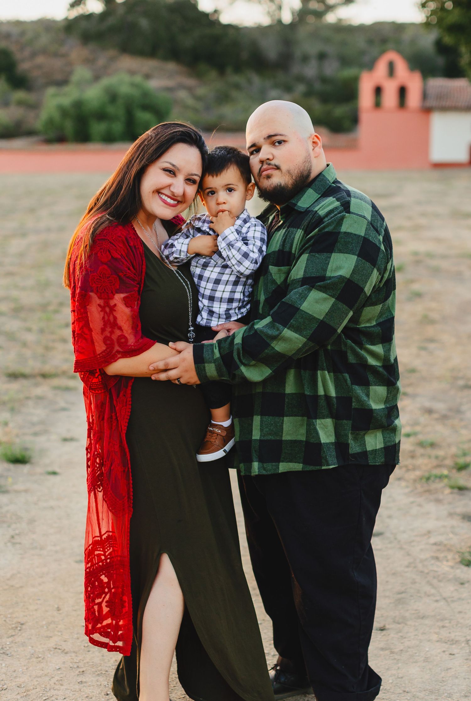 Family of three in casual attire poses together near a terracotta building at sunset.