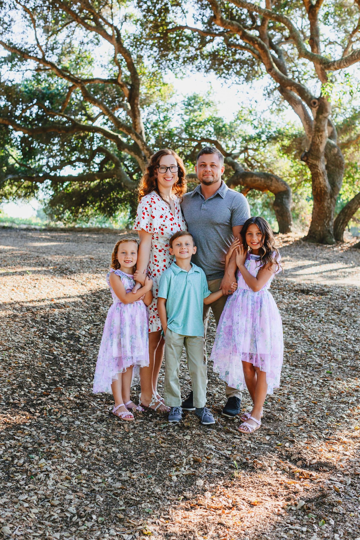 Family of five poses together under large oak trees in dappled sunlight.
