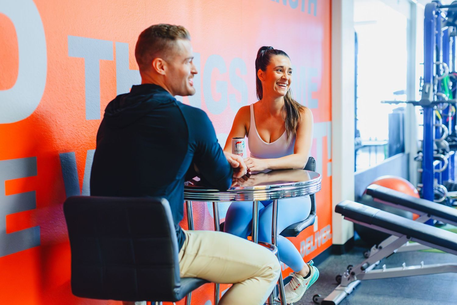 Two people having a conversation in modern orange and blue gym setting.